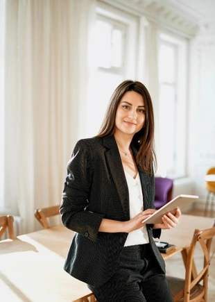 A professional woman in a business suit holding a tablet, standing confidently in a modern, well-lit office space.